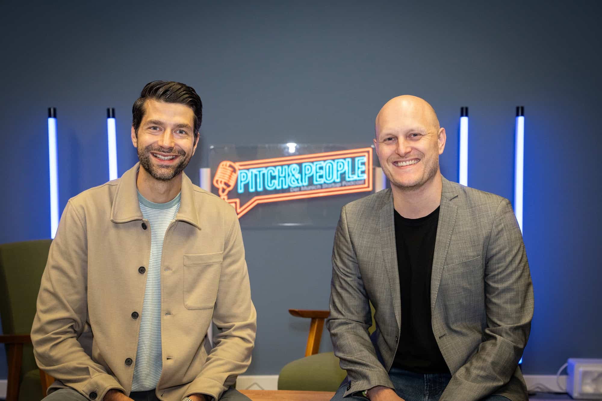 Zwei Personen im Pitch & People Podcast-Studio vor dem leuchtenden Podcast-Logo